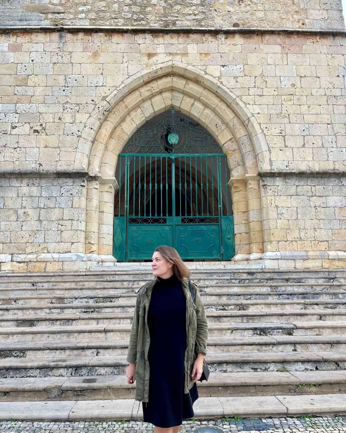 A woman in a black dress and a green jacket descends ancient stone steps in front of an imposing church door with intricate green metalwork. The setting evokes a sense of history and exploration, characteristic of solo travel adventures.