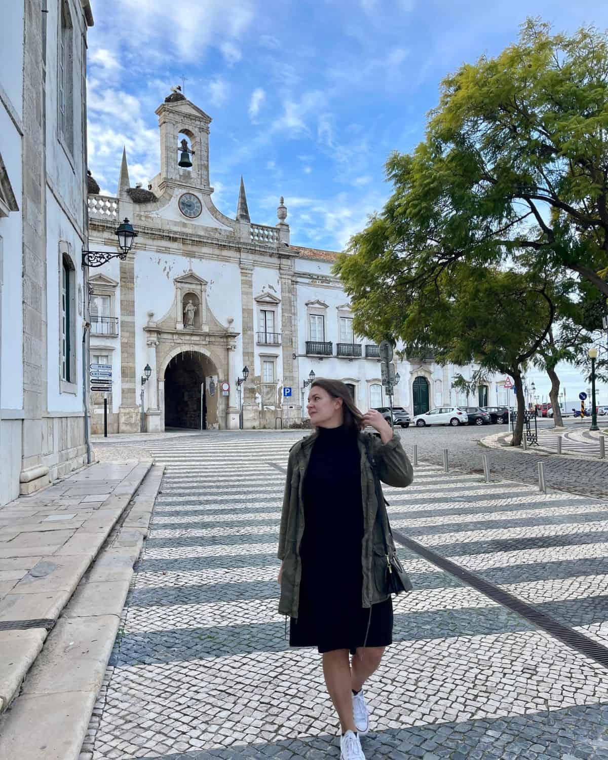 A solo female traveler in a black dress and green jacket stands on a cobblestone street in Faro, Portugal.