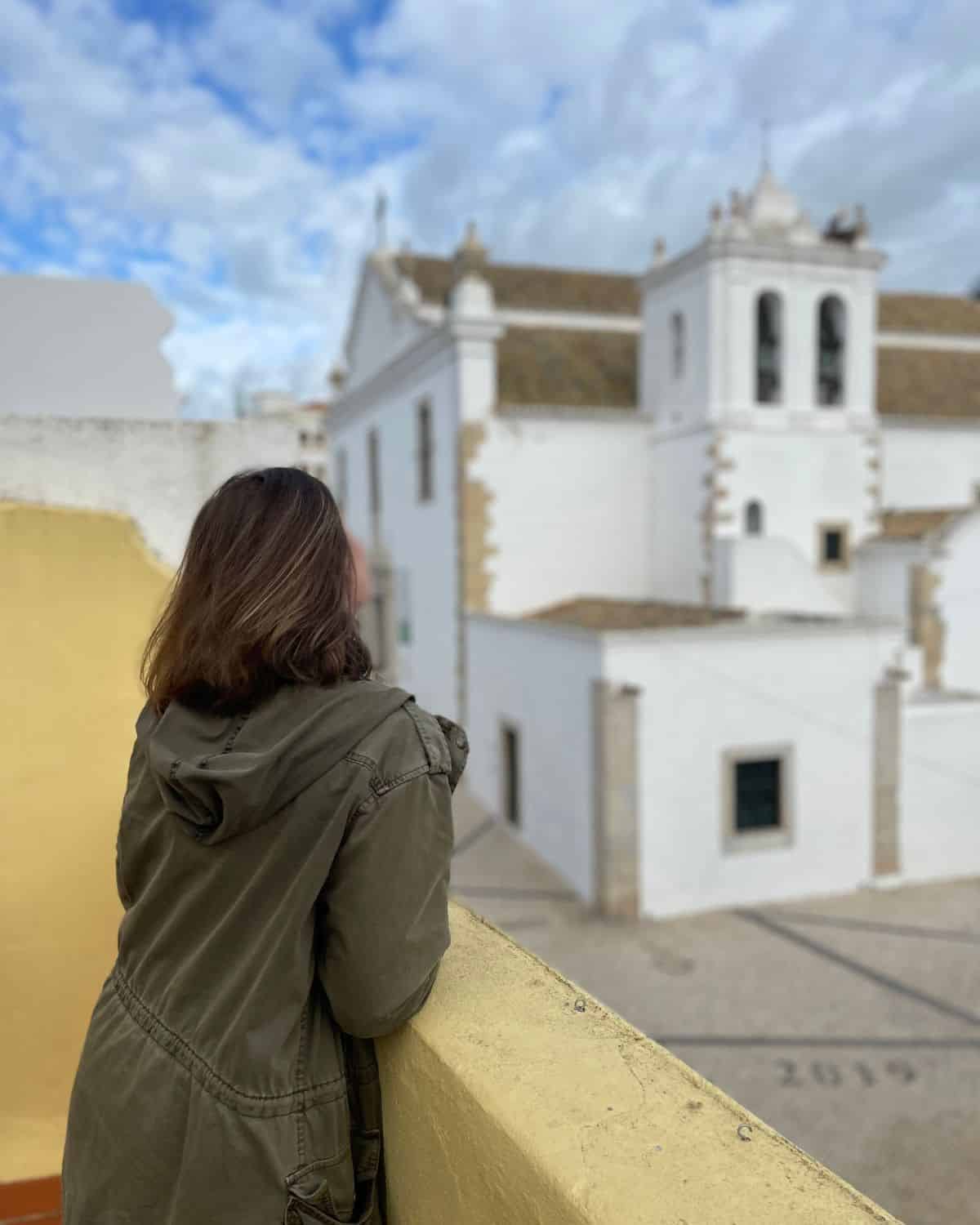View from behind the traveler as she leans on a yellow wall, looking out towards a quaint Faro neighborhood with traditional white buildings and a church with two bell towers, under a bright blue sky with scattered clouds.