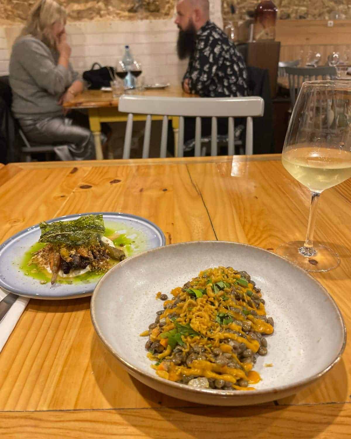 Image of a cozy restaurant interior with two plates of food in the foreground. The closest dish features a hearty, bean-based meal topped with crispy onions, and the other plate appears to have grilled fish on a bed of greens with a drizzle of sauce. In the background, a couple is engaged in conversation at their table. A glass of white wine is positioned to the right, complementing the dining experience.