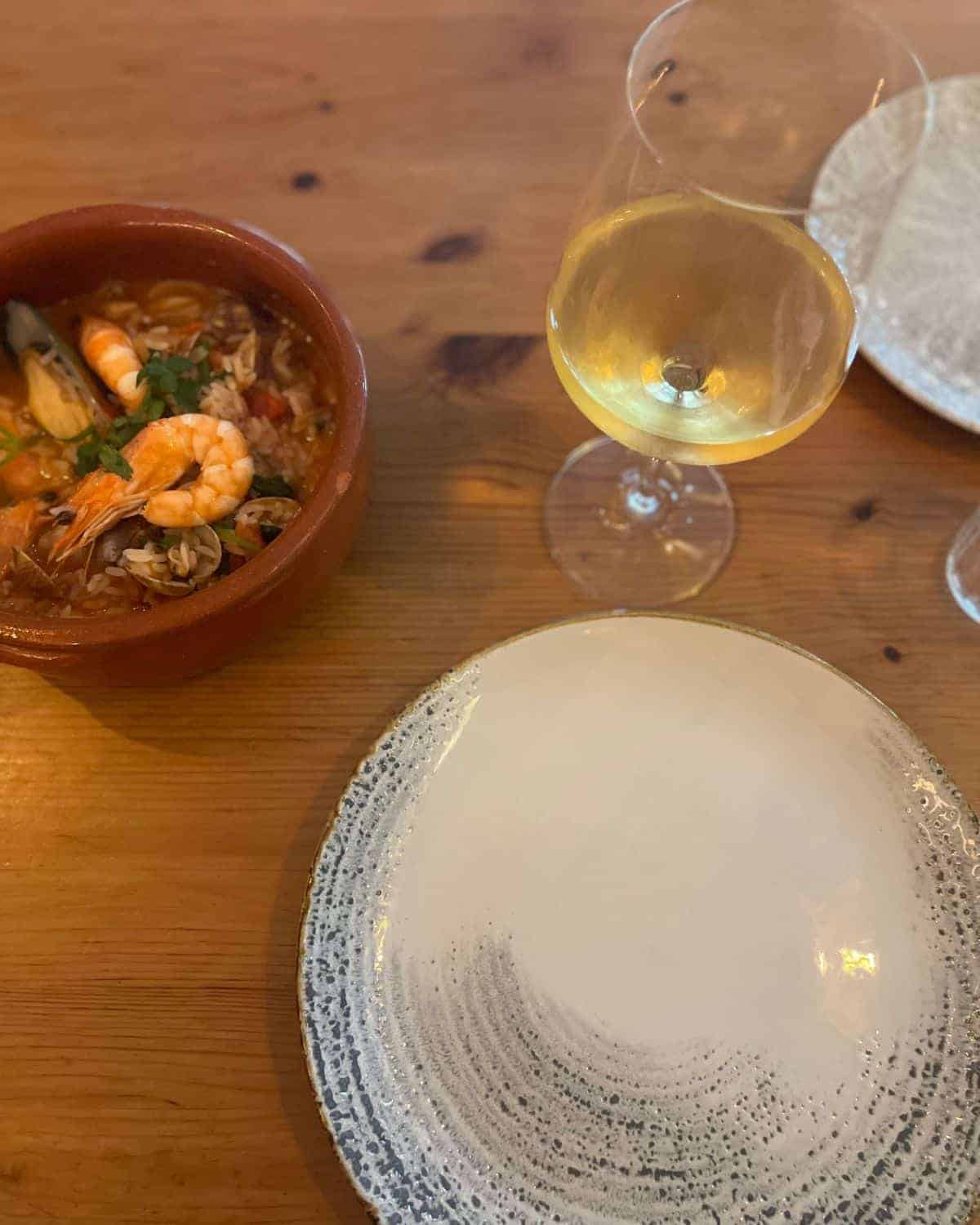 This image showcases a traditional dining setting with a terracotta bowl filled with what seems to be a seafood stew, rich in shrimps and clams, possibly a local delicacy. The bowl is paired with a glass of white wine, and another empty plate waits to be served, suggesting a meal shared.