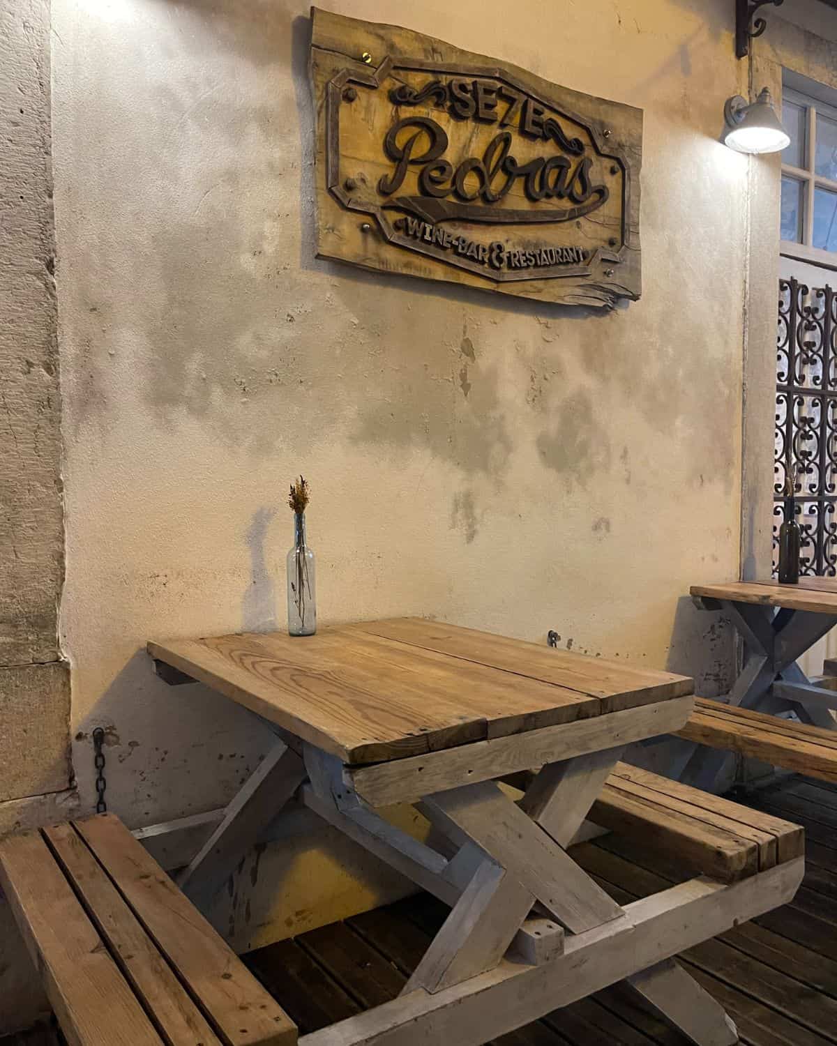 An outdoor seating area of a restaurant, featuring a wooden picnic-style table and benches. The rustic charm is accentuated by the textured, cream-colored wall and a vintage wooden sign reading "Taberna Pedras", indicating the establishment's name.