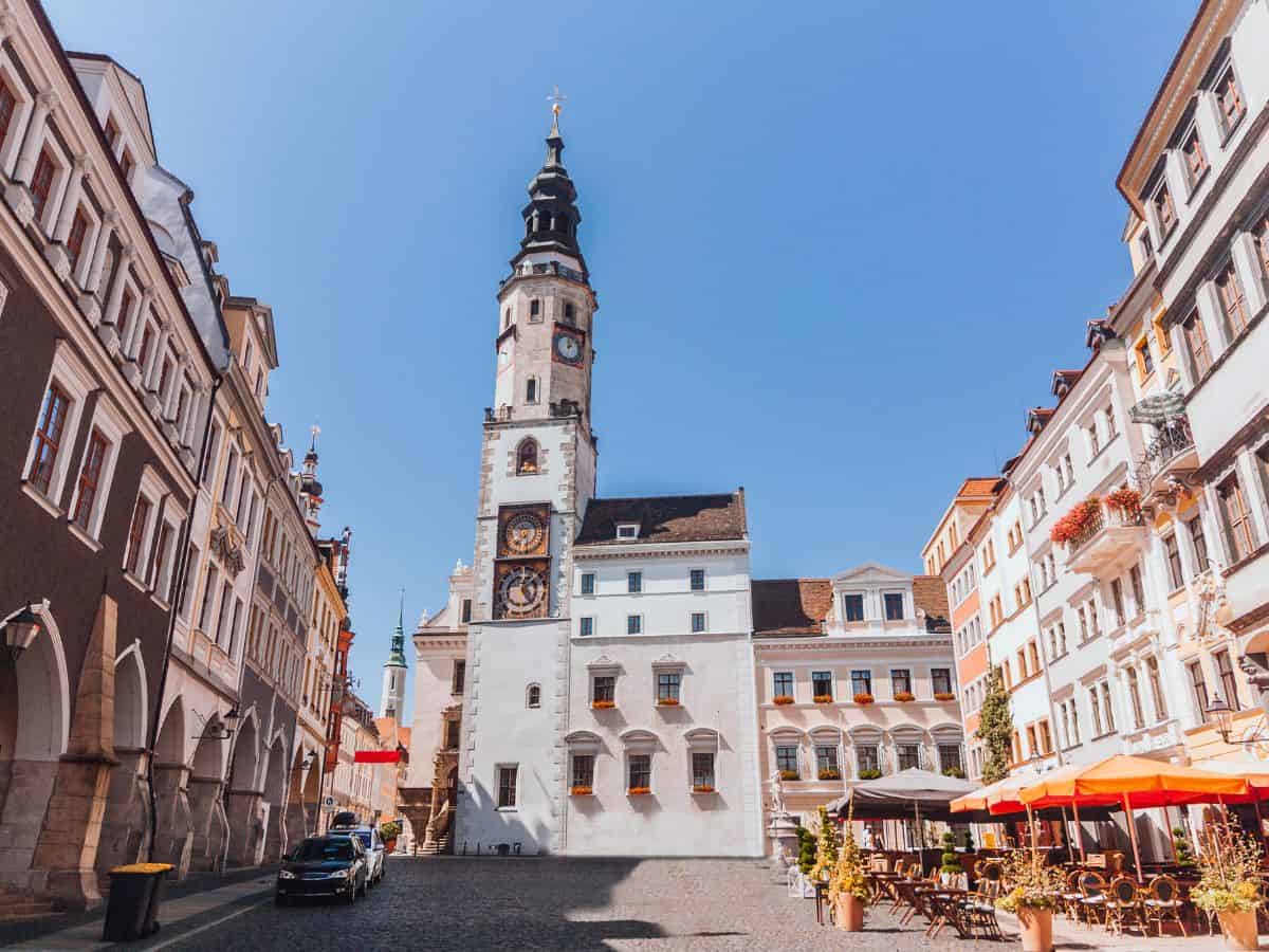 A quaint European square lined with colorful, traditional buildings under a clear blue sky. The centerpiece is an ornate historical tower with a clock, accented by its spiraling black and white patterned spire. To the right, an inviting café terrace awaits with its orange umbrellas and arranged seating, offering a perfect spot to relax and enjoy the serene atmosphere of Görlitz, a hidden gem of Europe.