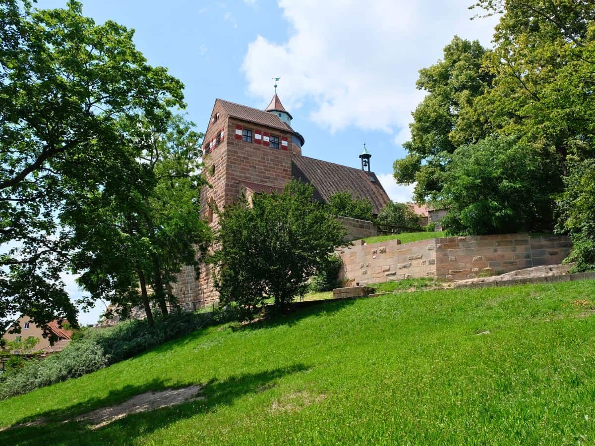 A sturdy tower stands atop a grassy hill, partially obscured by the lush foliage of mature trees. The tower, made of sandstone, features a conical roof and small windows, reflecting the city's medieval architecture. The clear blue sky with soft clouds provides a tranquil backdrop for this historic structure.