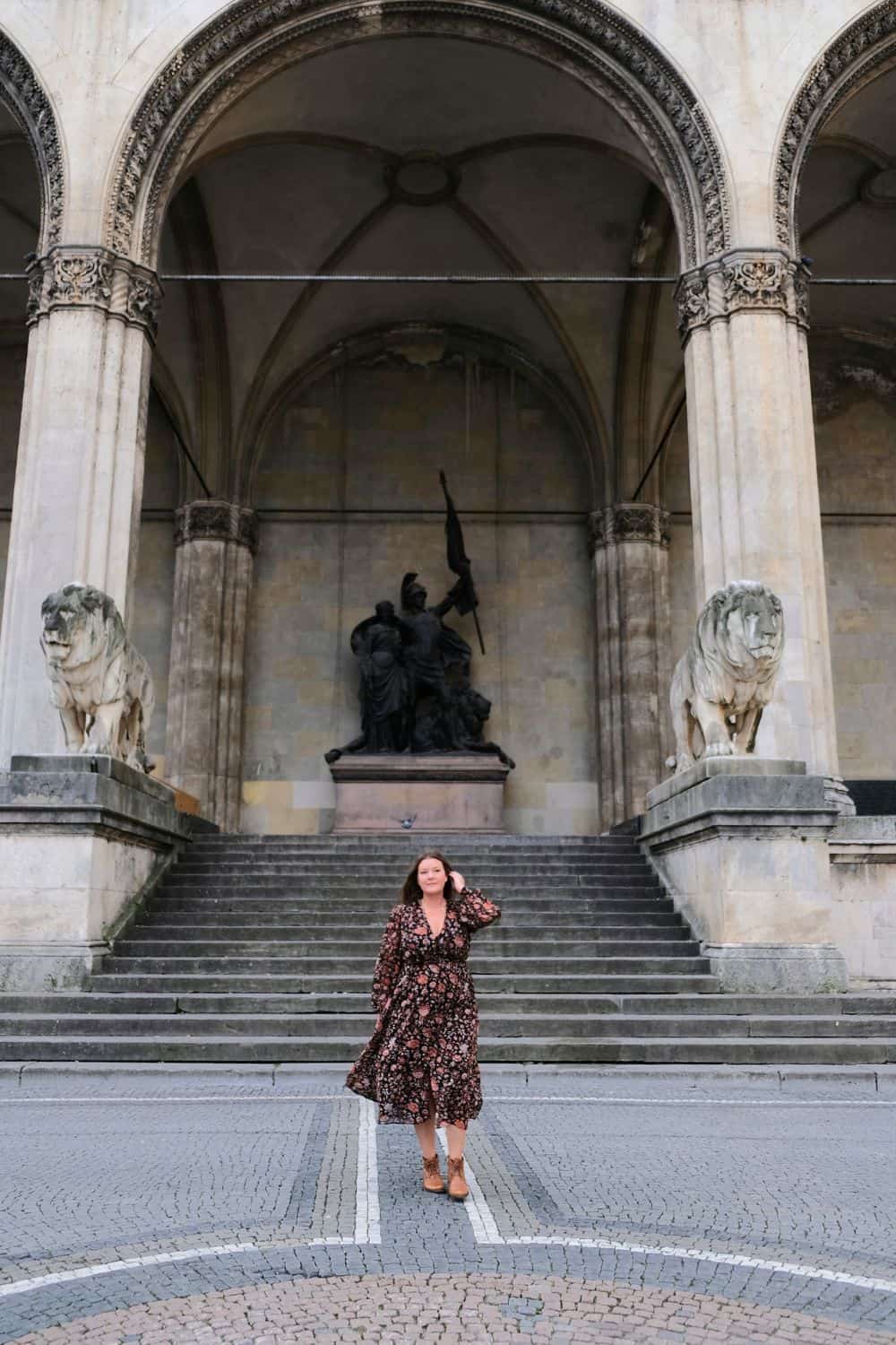 A woman in a floral dress stands before the majestic Feldherrnhalle in Munich, framed by historic statues and architecture, an iconic site for visitors.