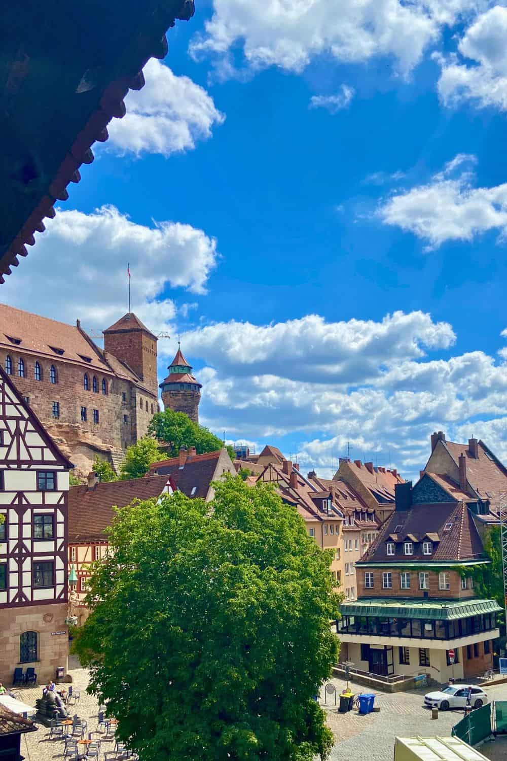A captivating view of Nuremberg, Germany, on a sunny day, showcasing the city's iconic medieval architecture. The image features half-timbered buildings and a sturdy stone castle towering over the old town. The vibrant greenery of trees contrasts with the quaint, cobblestone streets and the clear blue sky punctuated by fluffy white clouds, inviting exploration and discovery in this historic city.