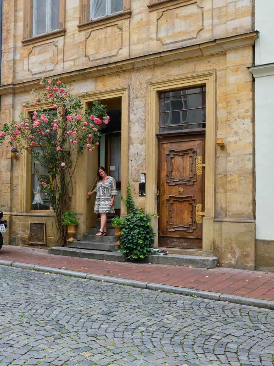 A female traveler smiles as she exits a vintage stone building adorned with blooming pink roses, capturing the essence of quaint streets ideal for day trips in the Nuremberg region