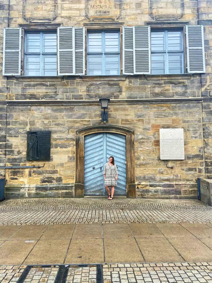 A woman stands centered in front of a grand stone doorway, with historical plaques adorning the wall in Bamberg