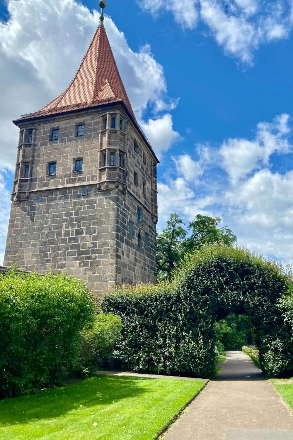 A vibrant view of a historic tower in Nuremberg, with its steep red roof and stone facade, rising majestically against a bright blue sky with wisps of white clouds. The tower is surrounded by lush greenery and a neatly trimmed hedge archway, leading to a path that promises an intimate glimpse into the city's medieval past.