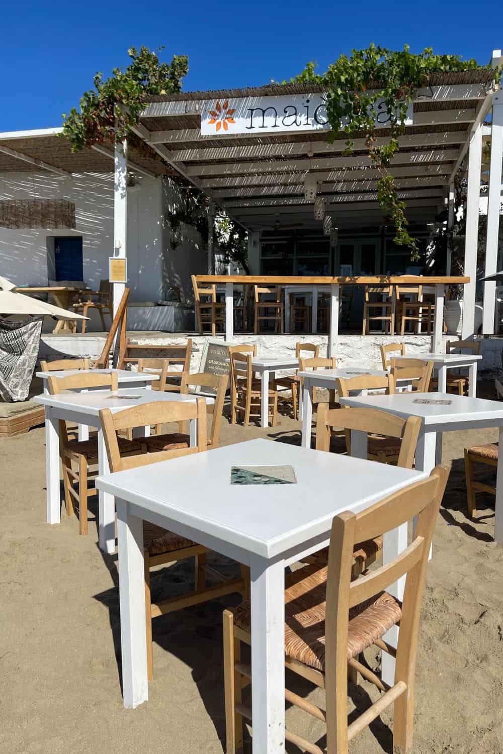 A beachside restaurant named 'Maiolica' basks in the sunlight, showcasing white tables with wooden chairs on sandy ground, ready to welcome diners with its charming, open-air ambiance.
