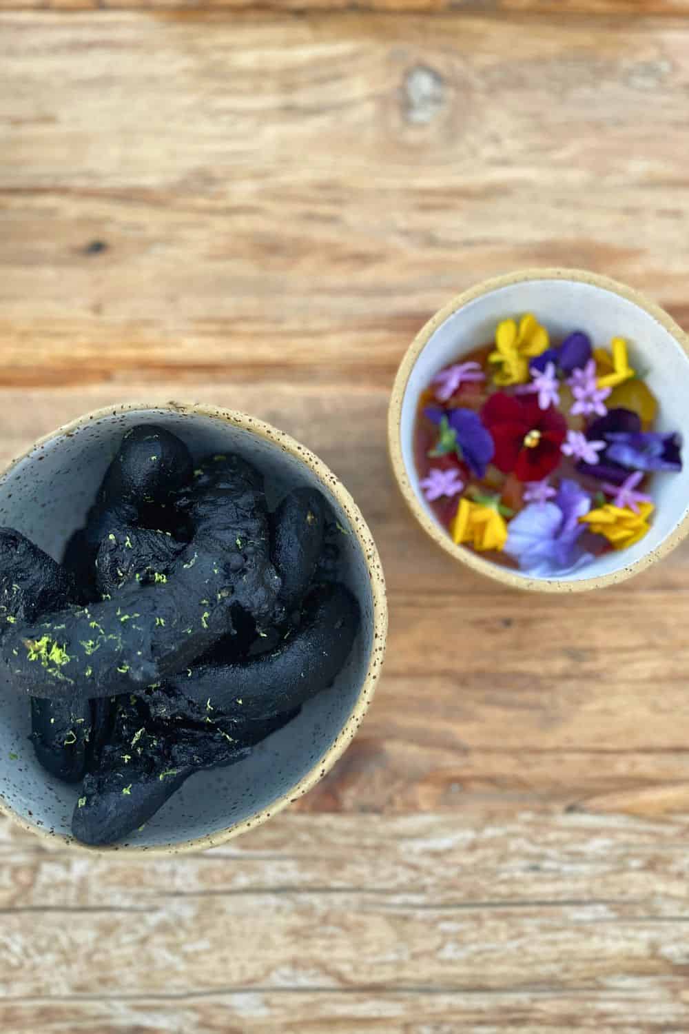 two bowls on a wooden surface, one containing charcoal-black bread, the other filled with a variety of colorful edible flowers in a clear broth, representing a contrast between dark and vividly colored culinary elements.
