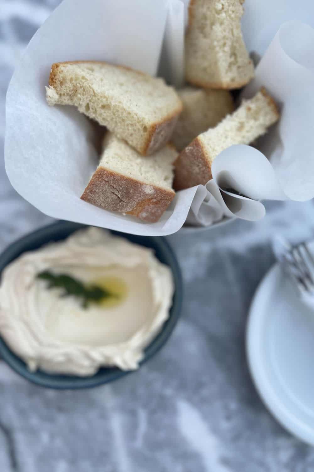 A casual dining setup featuring a paper-lined basket of freshly sliced bread with a side of creamy spread topped with olive oil and a single green leaf, presented on a marble table with a soft-focus background