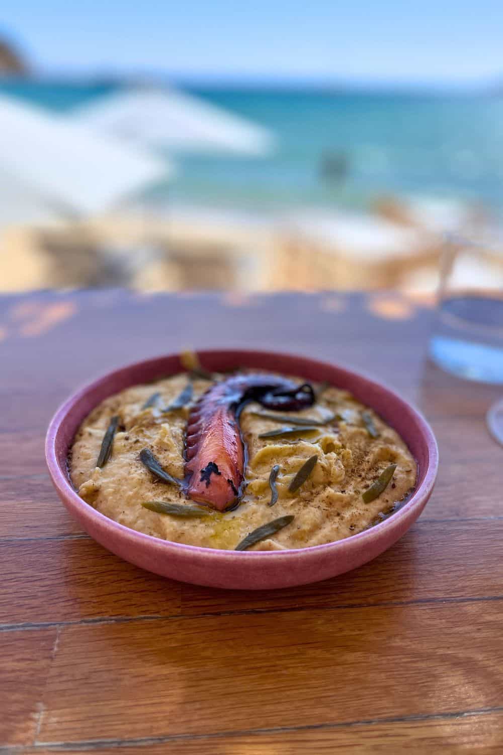 A close-up of a rustic, terracotta bowl filled with a creamy dip topped with a grilled pepper, set against a blurred backdrop of a sunlit beach and azure waters.