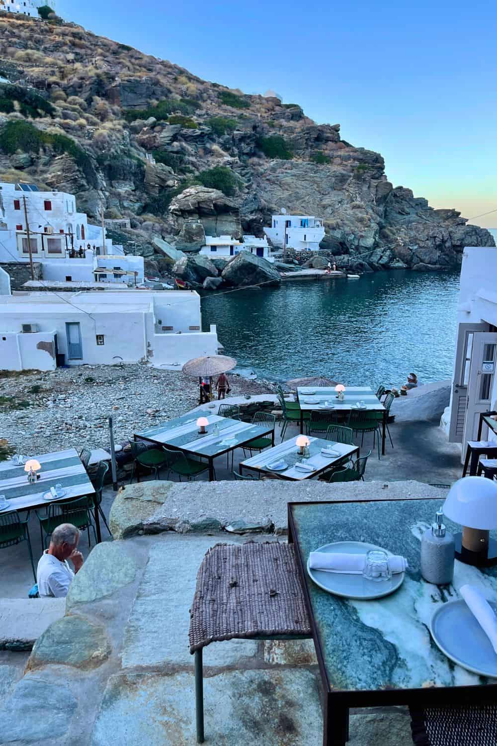 An evening dining setting by the sea in a quaint village, with green tables on a rocky shoreline, overlooking tranquil waters and white-washed buildings nestled against rugged cliffs.