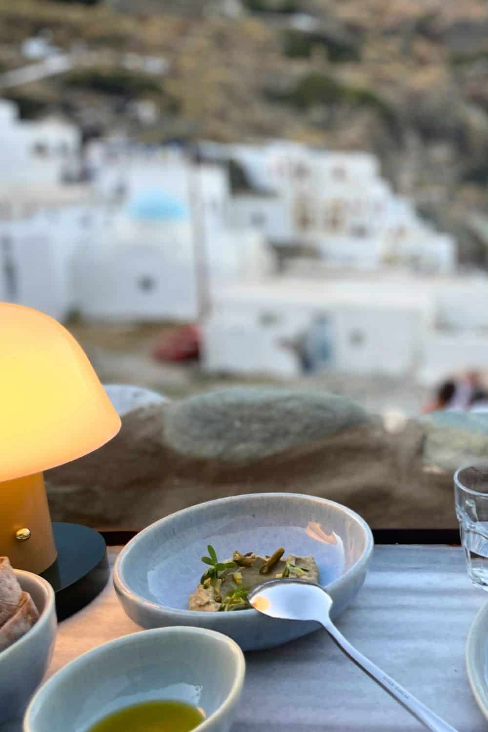 A serene dining table view at dusk, featuring a small bowl with a light appetizer, a dimly lit lamp, and overlooking a picturesque village descending towards the sea.