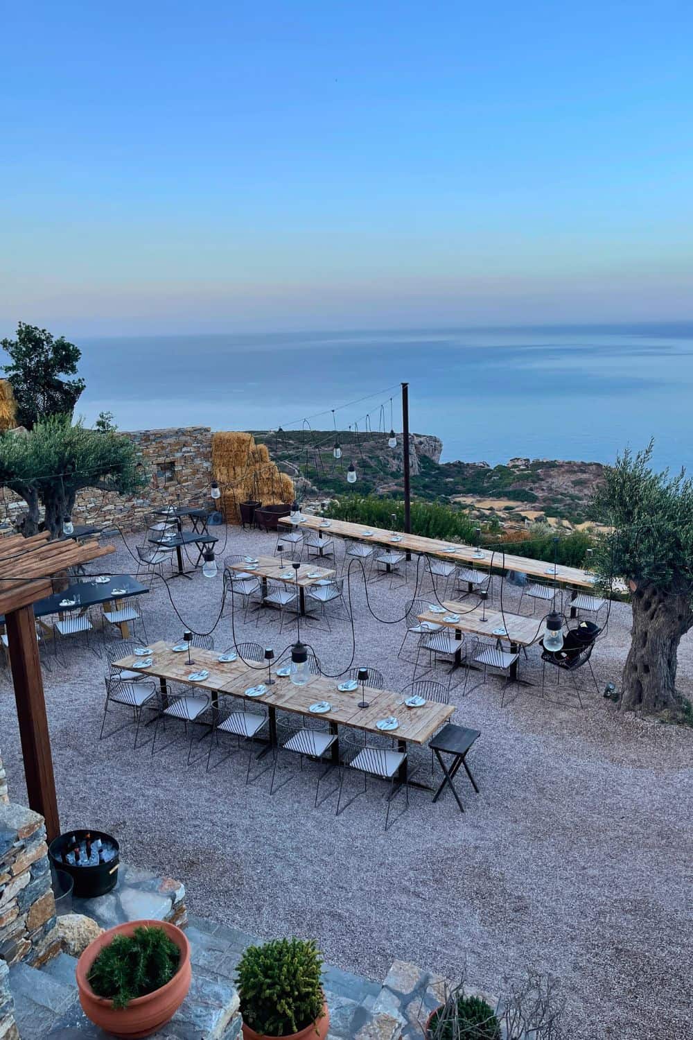 An open-air restaurant set on a hill, with an expansive view of the sea at twilight, featuring rows of simple yet elegant tables and chairs spread out on a gravel-covered terrace