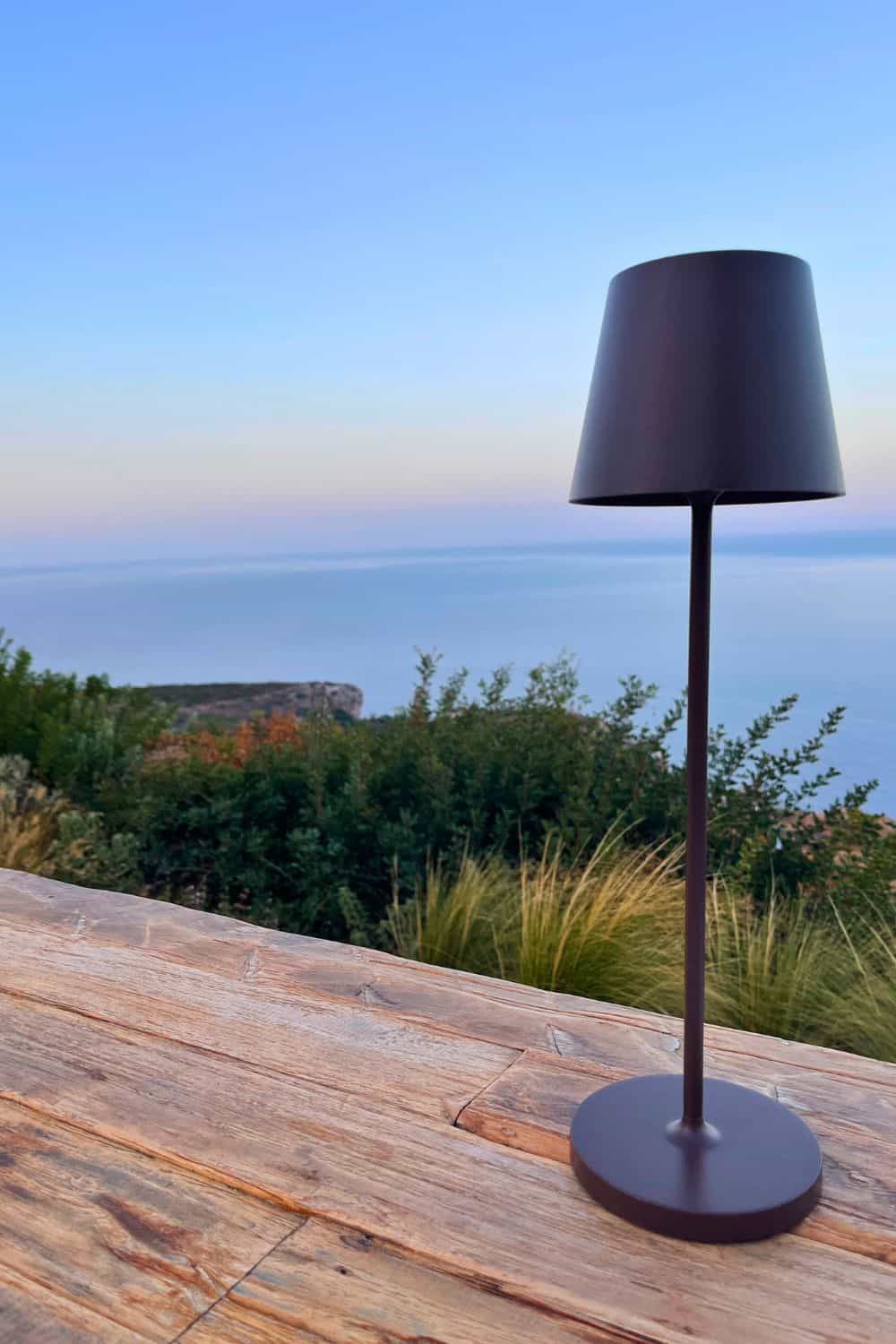 A modern lamp stands on a wooden table, with a panoramic view of the twilight sky meeting the calm sea, conveying a peaceful dining experience on a hillside.