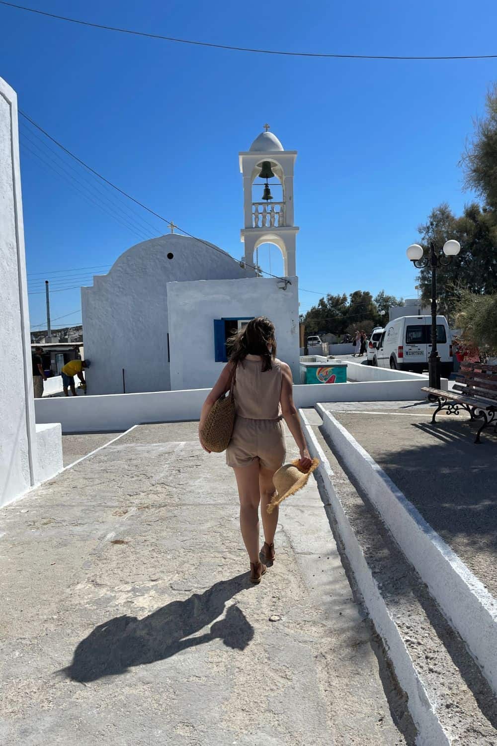 A woman walking towards a traditional Greek church with a bell tower, on a sunny day in Milos