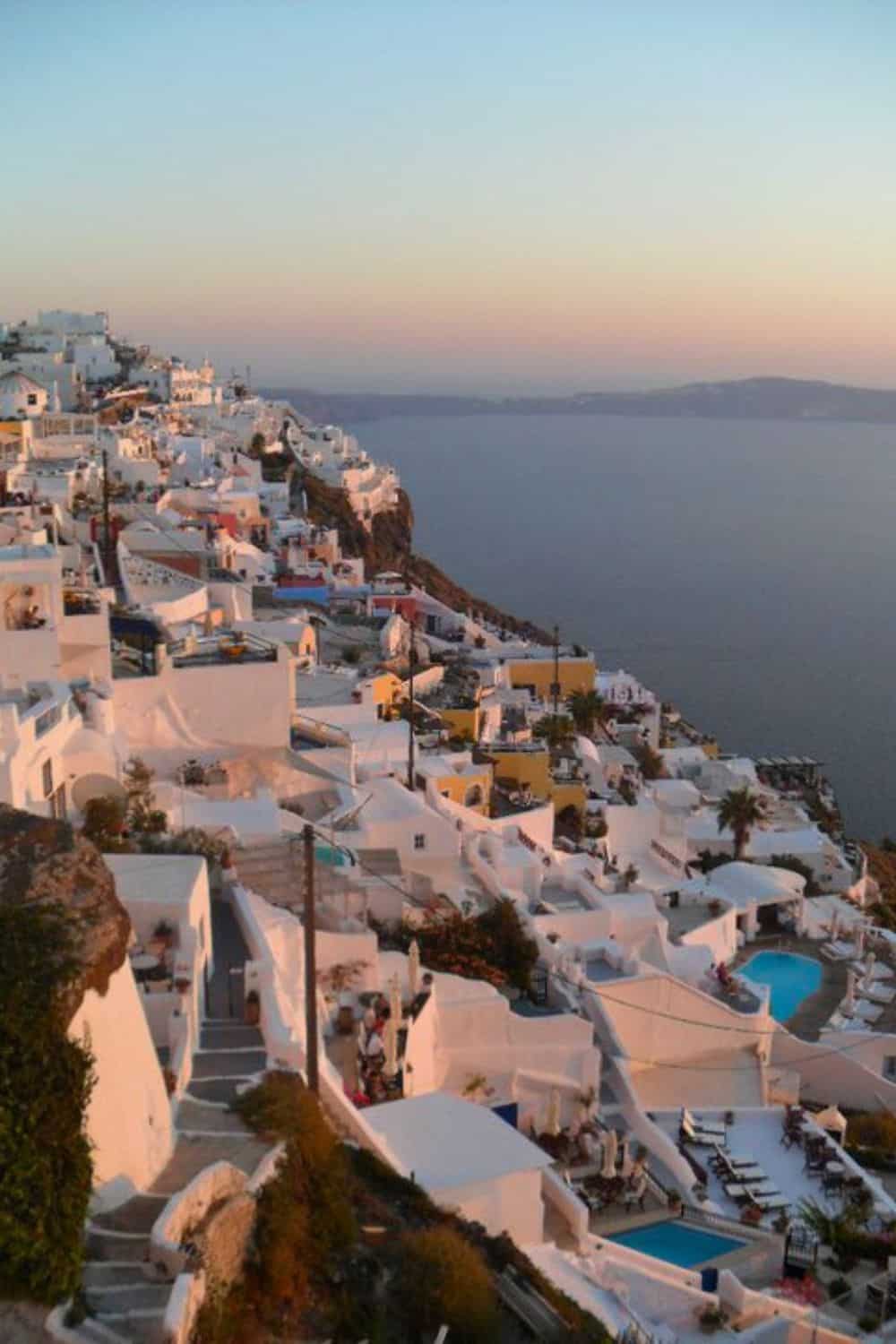 Twilight over Santorini: A cascade of traditional white Cycladic houses with blue accents descends the cliffside toward the Aegean Sea, with the sky softly transitioning from pink to blue at dusk.