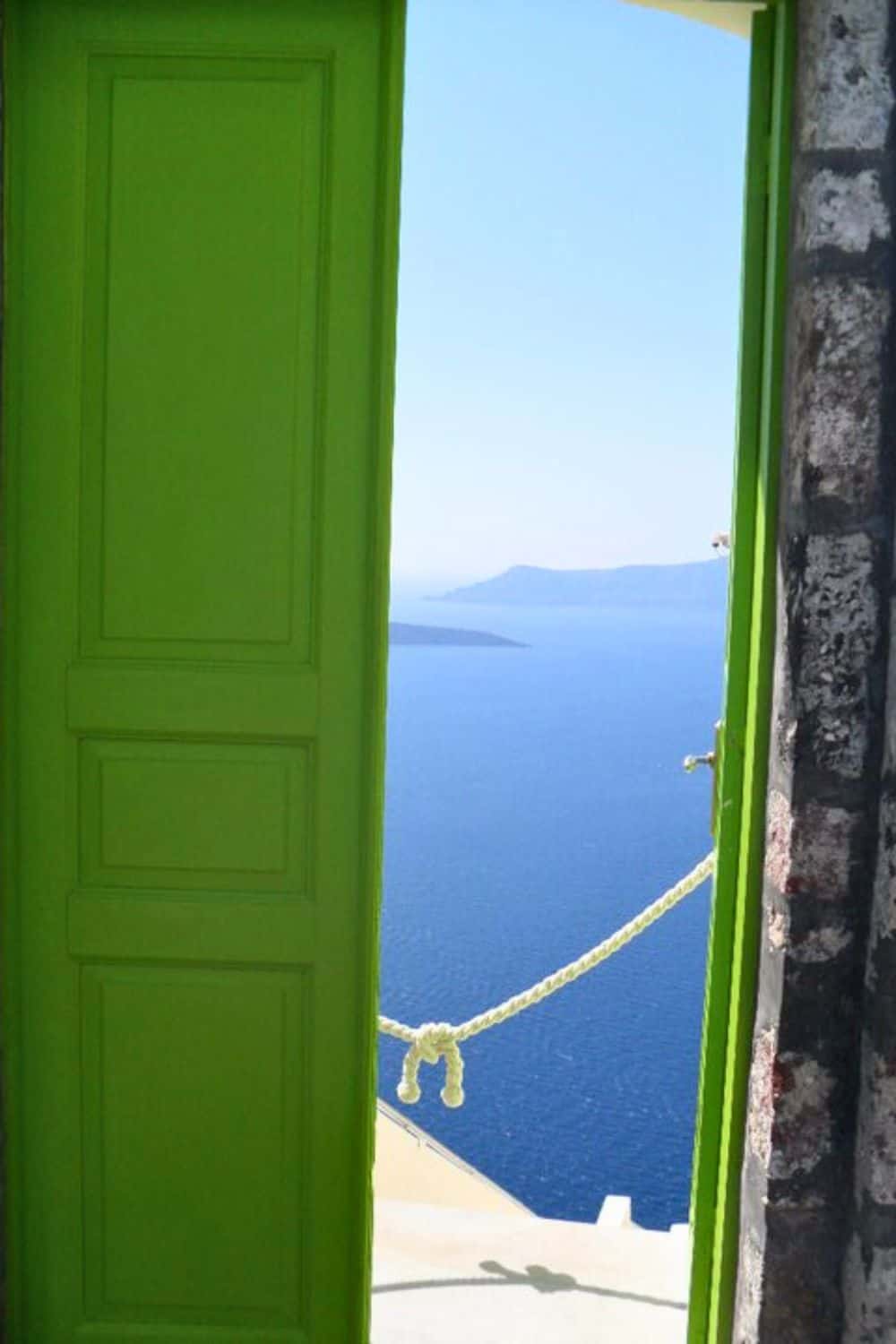 A vivid green door opens to reveal a stunning view of the bright blue Aegean Sea, with a simple rope across the entrance suggesting an invitation to the serene seascape beyond.