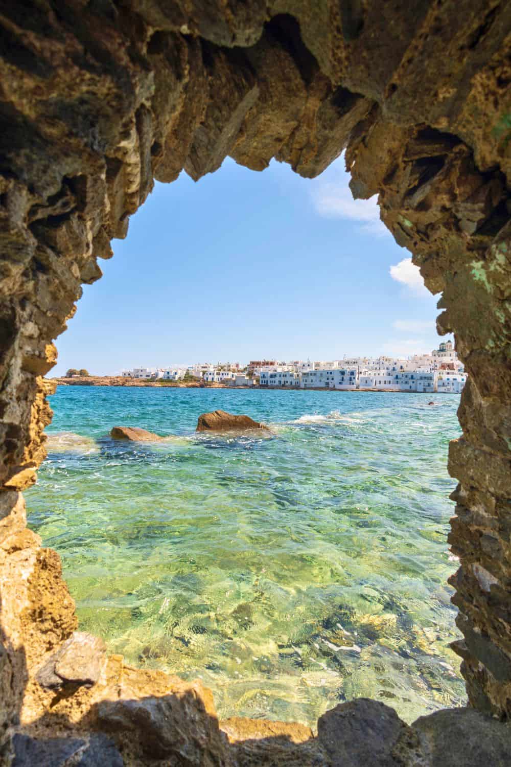 View from a rocky cave on Paros, framing the tranquil azure waters of the Aegean Sea with the white-washed buildings of a distant coastal town under a clear blue sky