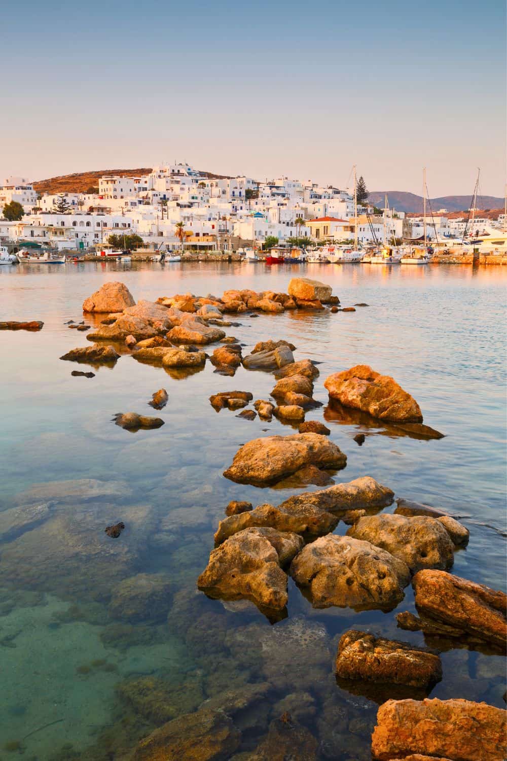 Golden hour at a Paros seaside: The warm glow of sunset illuminates the rocky shore and the calm sea, with the traditional whitewashed town in the background reflecting the last sunbeams.