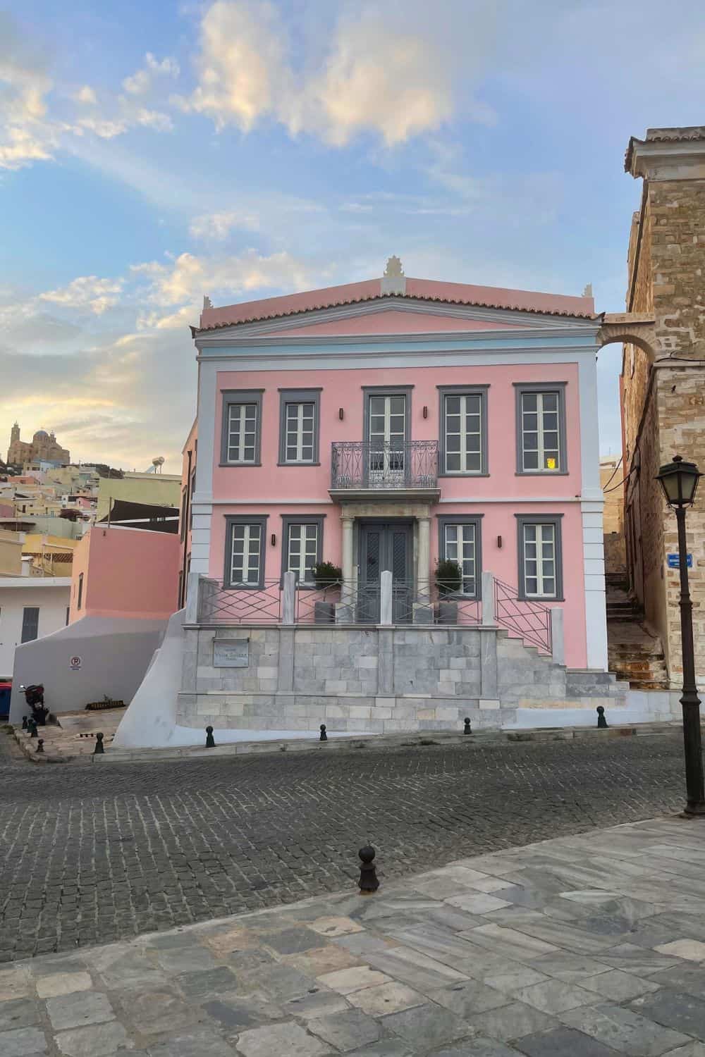 A charming, two-story pink neoclassical building with contrasting grey stone foundation and ornate metal balcony railings, located on a cobblestone street corner in a historic neighborhood, with a clear blue sky above.