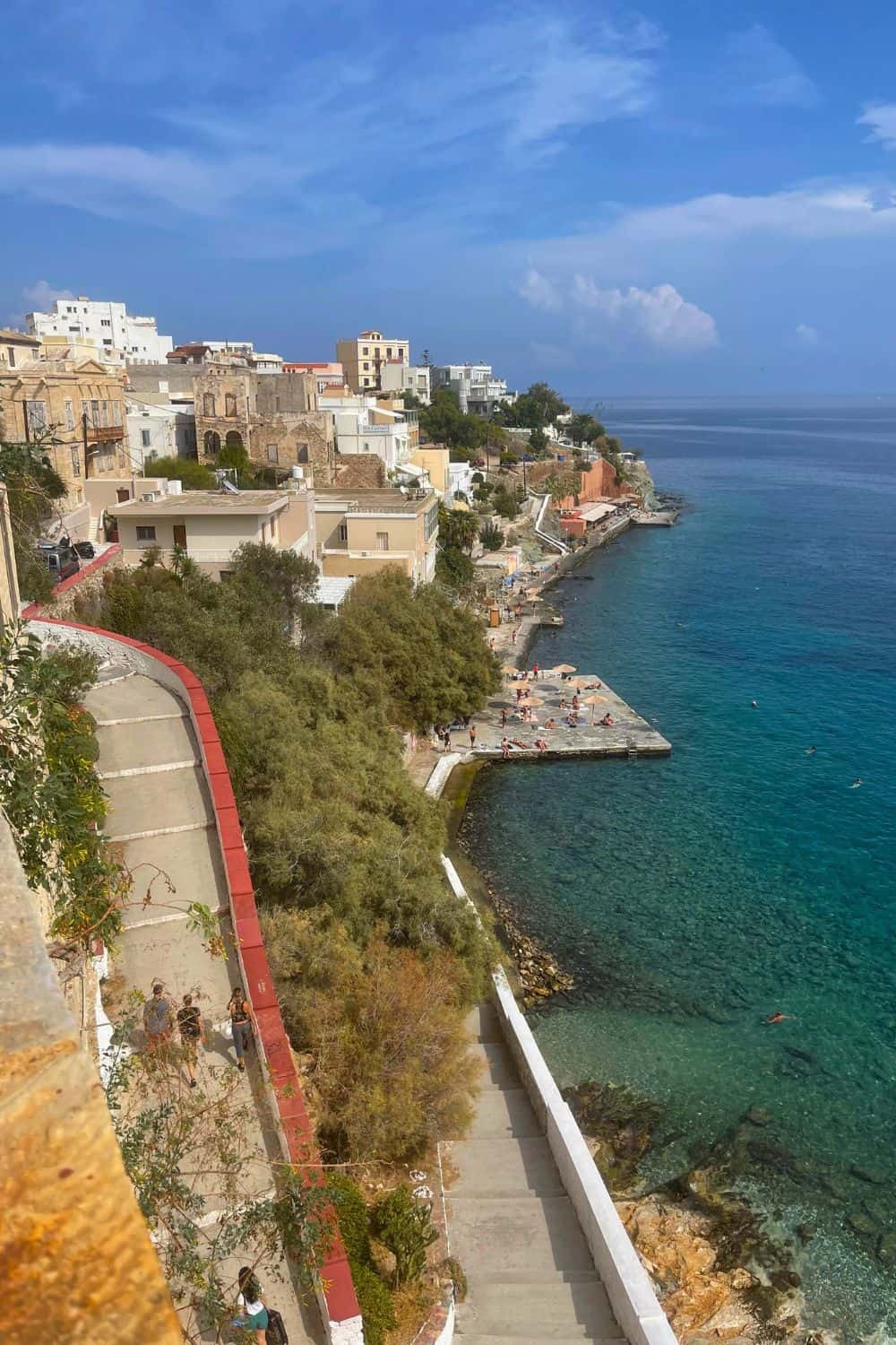 A coastal view from above showing a curving seaside promenade in Syros, with houses built along the cliff edge overlooking the clear blue waters of the Aegean Sea, and sunbathers enjoying the narrow beach below