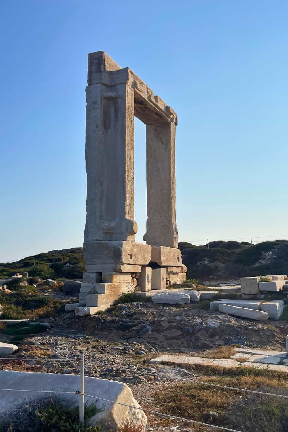 The Portara, a large marble doorway standing on a hill, remnants of an ancient temple on the island of Naxos, with clear blue sky in the background, and the sunlight casting a warm glow on the stone