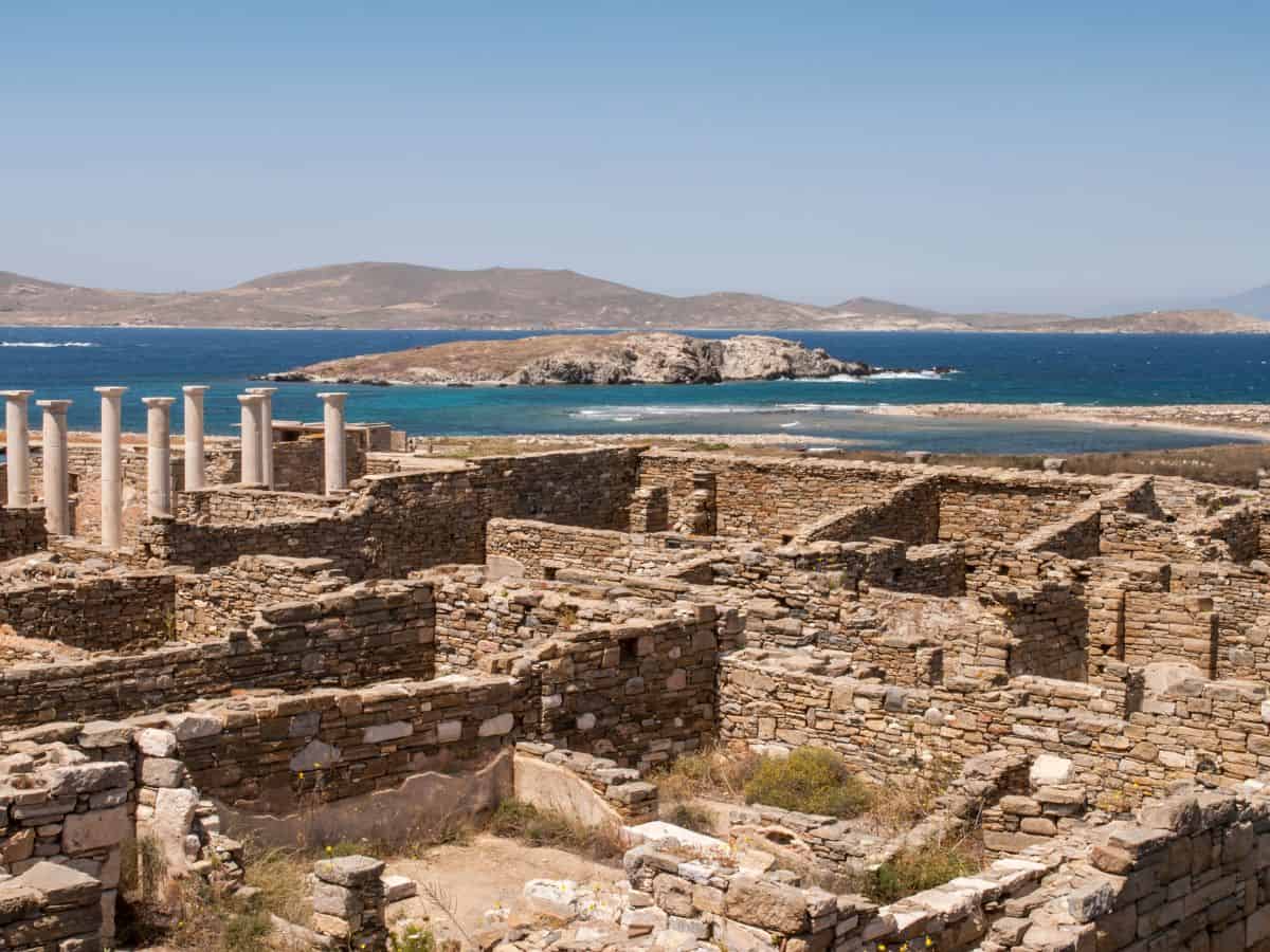 Ancient ruins on the island of Delos, Greece, with a row of intact marble columns standing in the foreground. Behind the archaeological remains, the landscape opens up to the blue sea and a rugged coastline under a clear sky