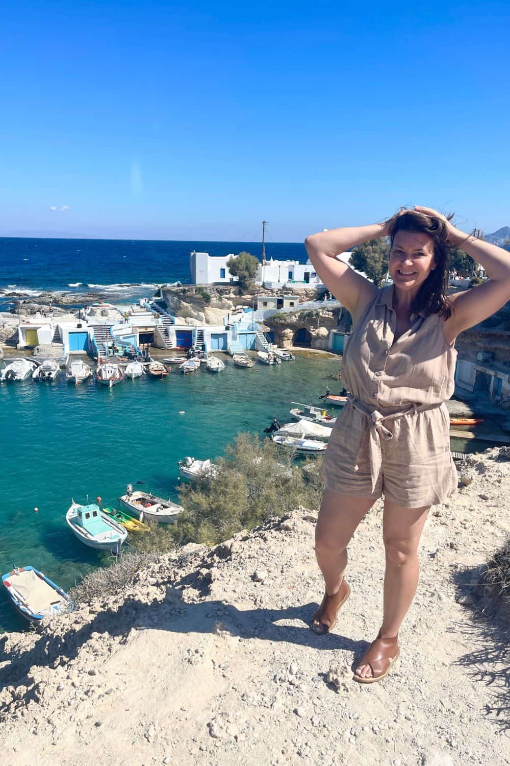 woman smiling at the camera, standing on a rocky outcrop with a panoramic view of a quaint Greek harbor with colorful boats in Milos.