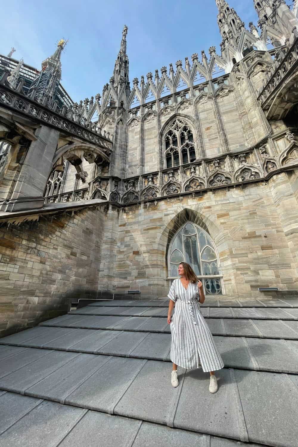 A woman in a striped dress stands on the rooftop of Milan Cathedral, with gothic spires rising behind her against a clear blue sky. She appears relaxed, gazing into the distance, capturing the grandeur of the historic architecture.