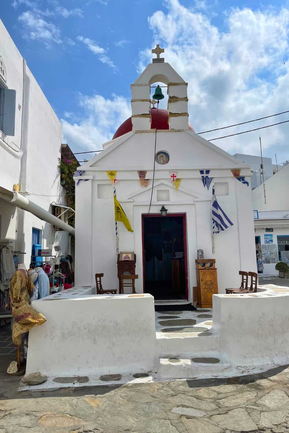 A small, charming church with a red dome and a white cross stands in the heart of Mykonos town. The façade is decorated with religious icons and Greek flags, while the steps leading up to the entrance are lined with chairs, showcasing the traditional Greek Orthodox architecture and community spirit of the island
