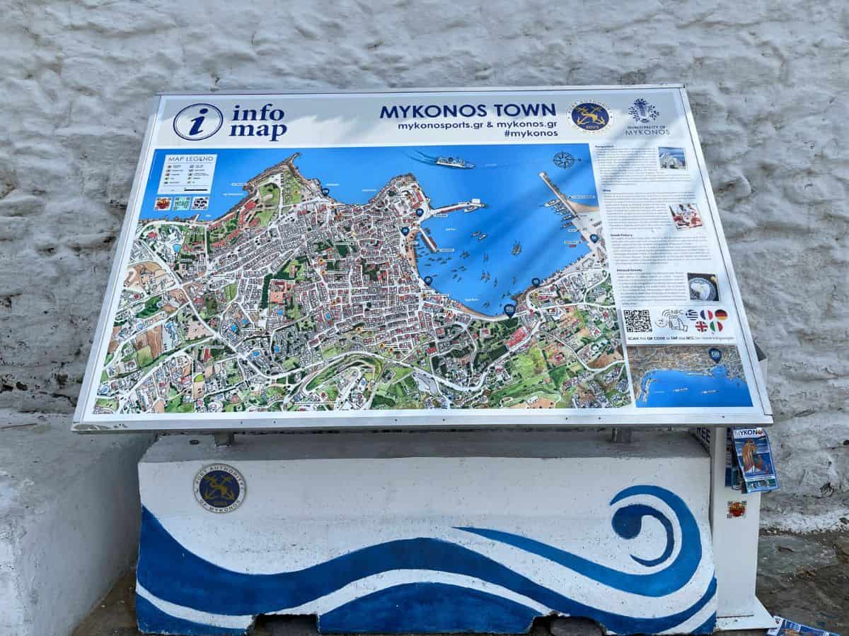 An informational map of Mykonos Town, featuring a colorful and detailed layout of the streets and key landmarks. The map includes a legend and various points of interest are marked. The map is mounted on a white and blue stand with a wave design, emblematic of the Greek flag colors.