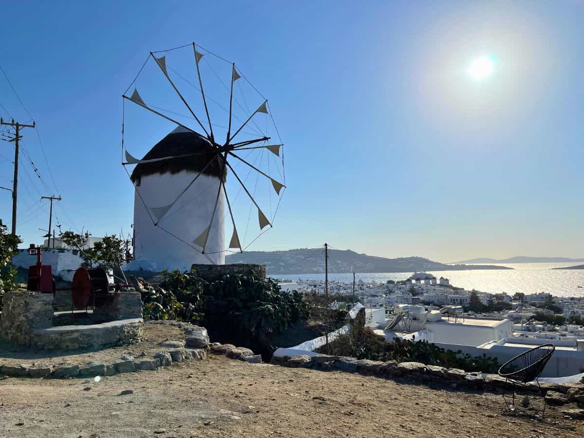 A traditional Greek windmill against a clear sky, with the sun shining brightly to the right. The windmill, with its characteristic white structure and thatched roof, is in the foreground. In the background, there's a panoramic view of Mykonos town, showing white buildings and the shimmering sea stretching to the horizon.