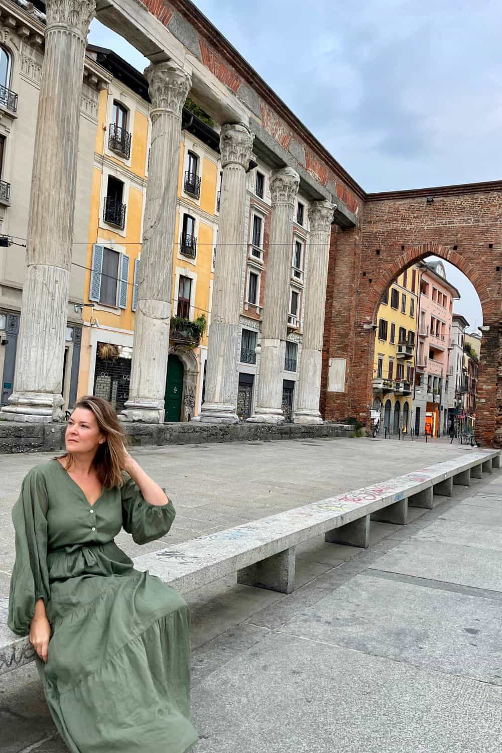 A woman in a green dress sits casually on a long stone bench in front of the ancient Columns of San Lorenzo in Milan. Behind her is a contrast of historic columns and vibrant modern buildings, embodying the blend of old and new in the city.