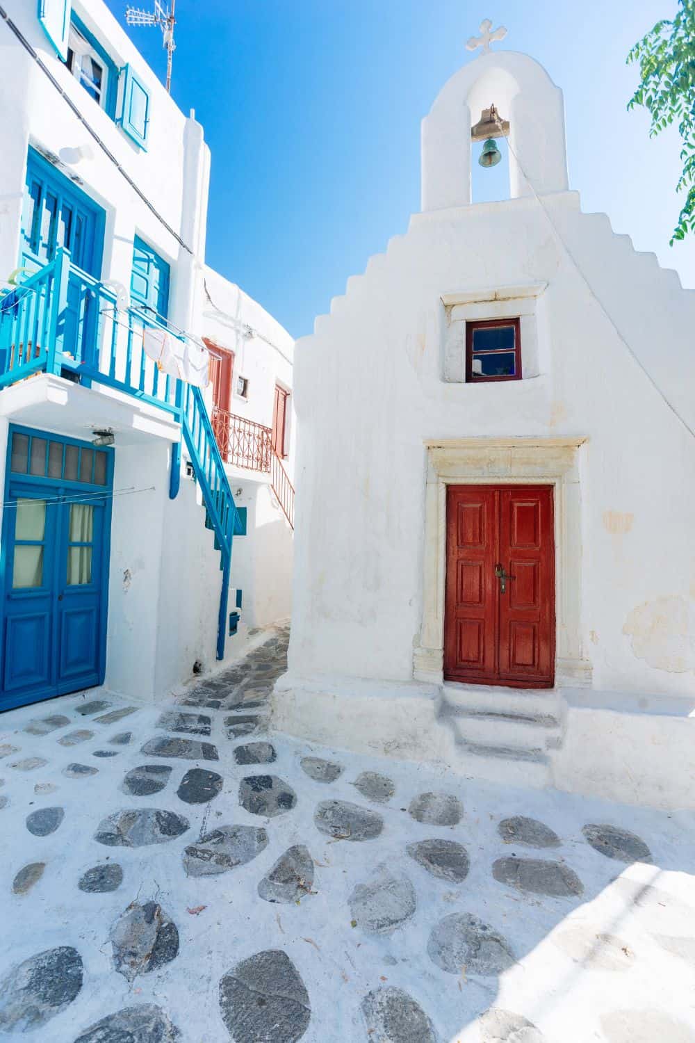 A charming narrow alley in Mykonos, with a white-washed church featuring a red door and blue-framed window. The cobbled path and bright blue and red accents on the buildings highlight the traditional Cycladic architecture.