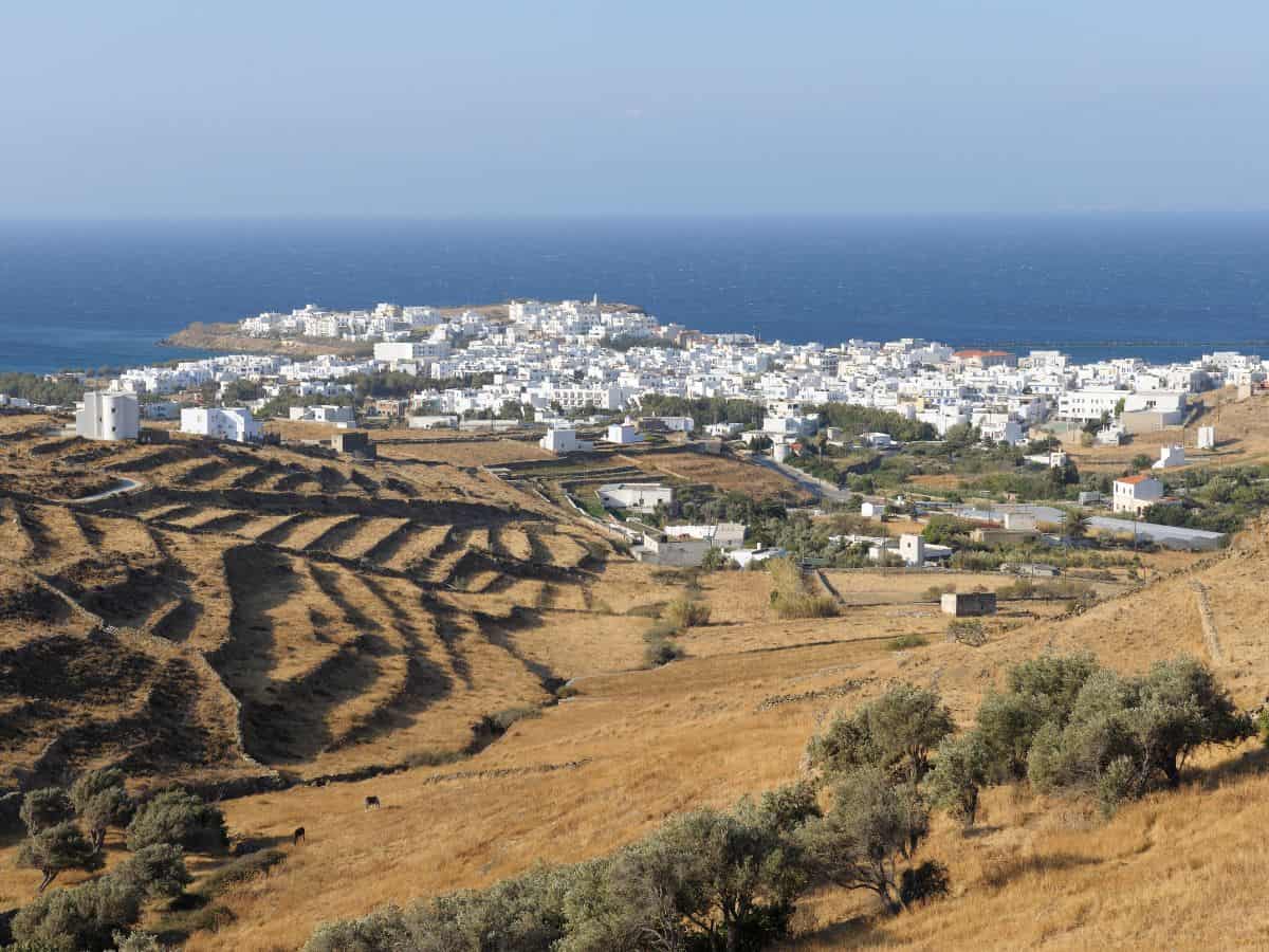 Panoramic view of the island of Tinos, Greece, featuring terraced fields on the rolling hills in the foreground, leading to the clustered white buildings of the town in the distance, with the Aegean Sea extending to the horizon