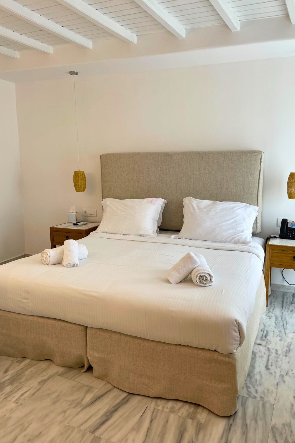 A neatly made bed with a large beige headboard in a hotel room in Mykonos. The room features white walls, a white ceiling with exposed wooden beams, and a hanging pendant light with a natural fiber weave. The clean and simple design offers a serene and inviting atmosphere.