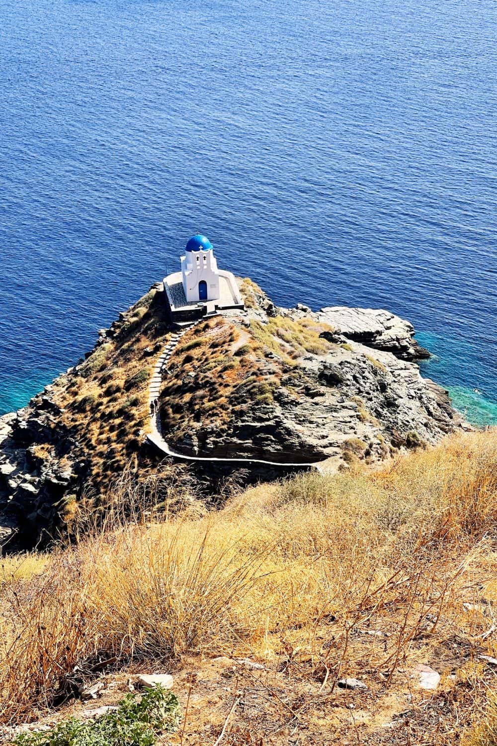 A solitary white church with a striking blue dome sits at the edge of a rocky promontory in Sifnos, overlooking the deep blue sea. The sun casts a brilliant light on the arid landscape, highlighting the rugged beauty of the island's coastline.