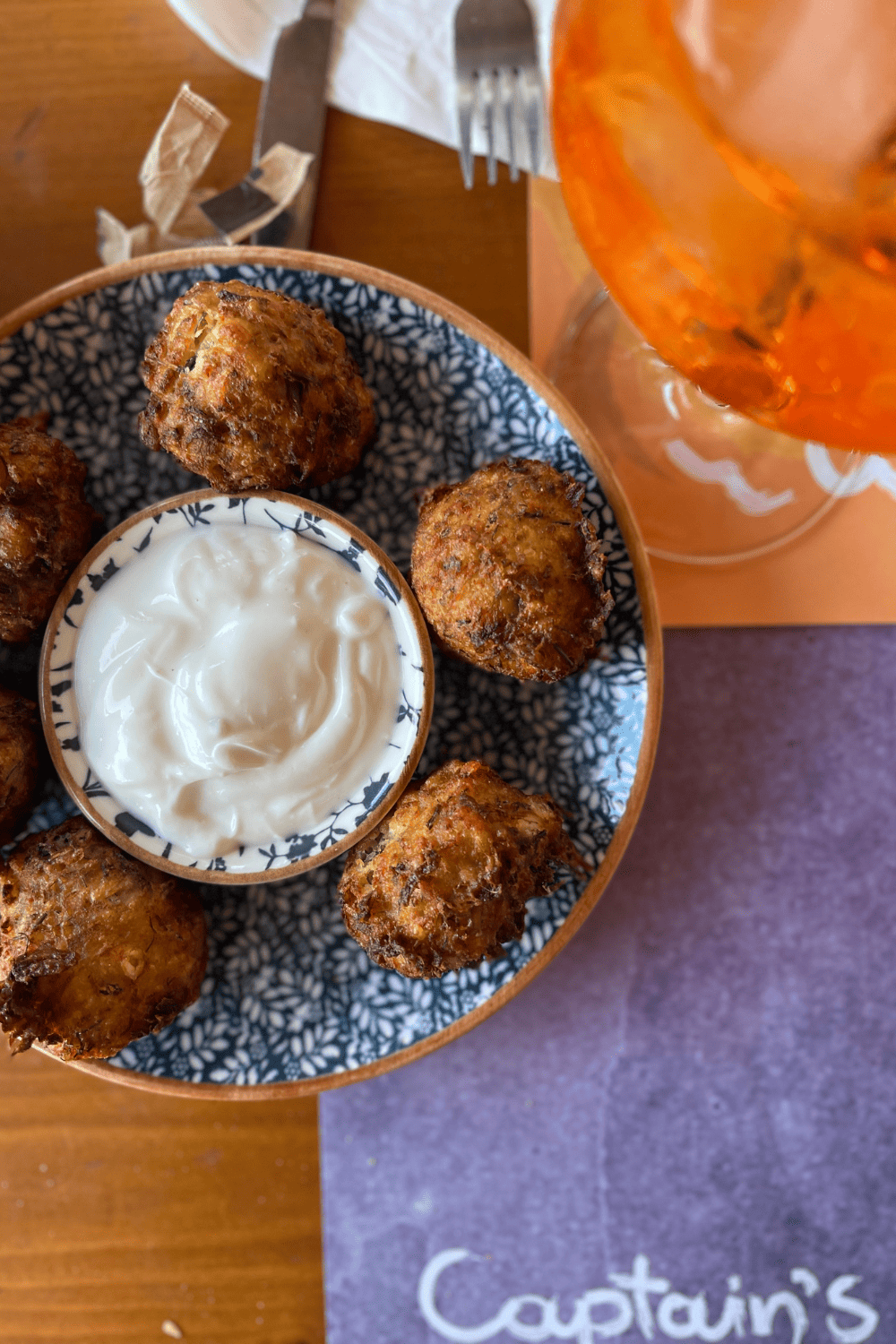 A close-up of a dish with fried zucchini balls served with a bowl of creamy tzatziki sauce on a decorative blue and white plate. An orange drink is blurred in the background, and the word 'Captain’s' is visible on a napkin, suggesting the setting is a restaurant.
