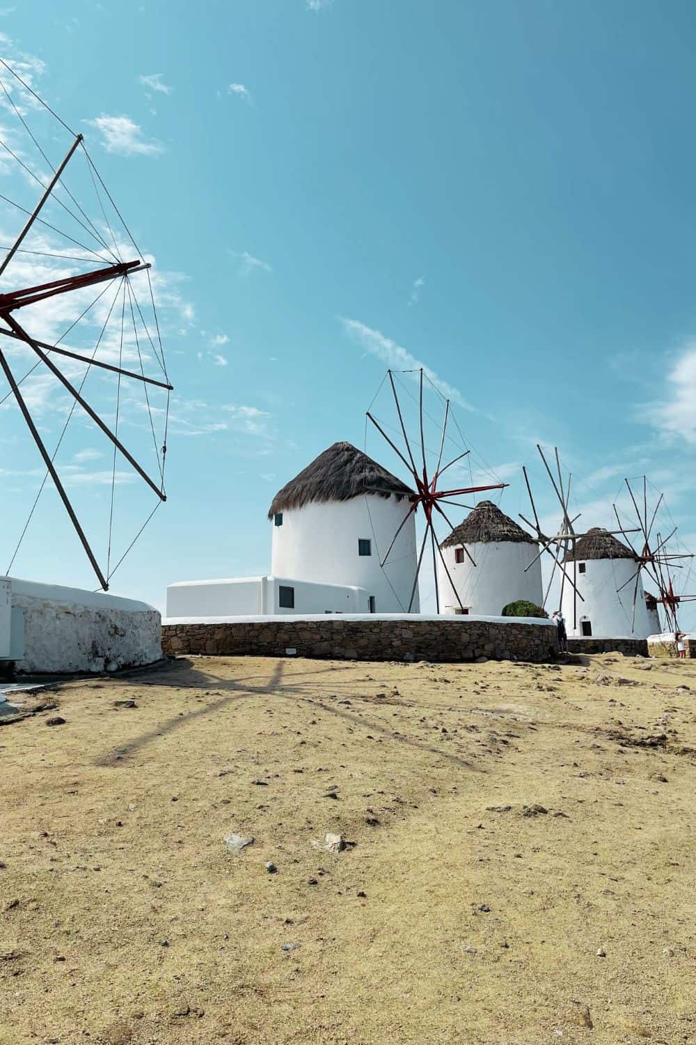 Iconic windmills of Mykonos against a clear blue sky. The foreground shows a barren landscape leading up to the famous white structures with thatched roofs and red-tipped wooden blades, characteristic of this Greek island.