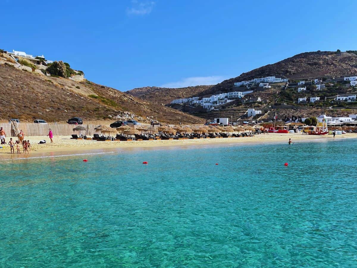 A beautiful beachfront view in Mykonos with crystal-clear turquoise waters and a sandy beach lined with sunbeds and umbrellas, leading to a backdrop of white buildings nestled against a rocky hillside under a clear blue sky.