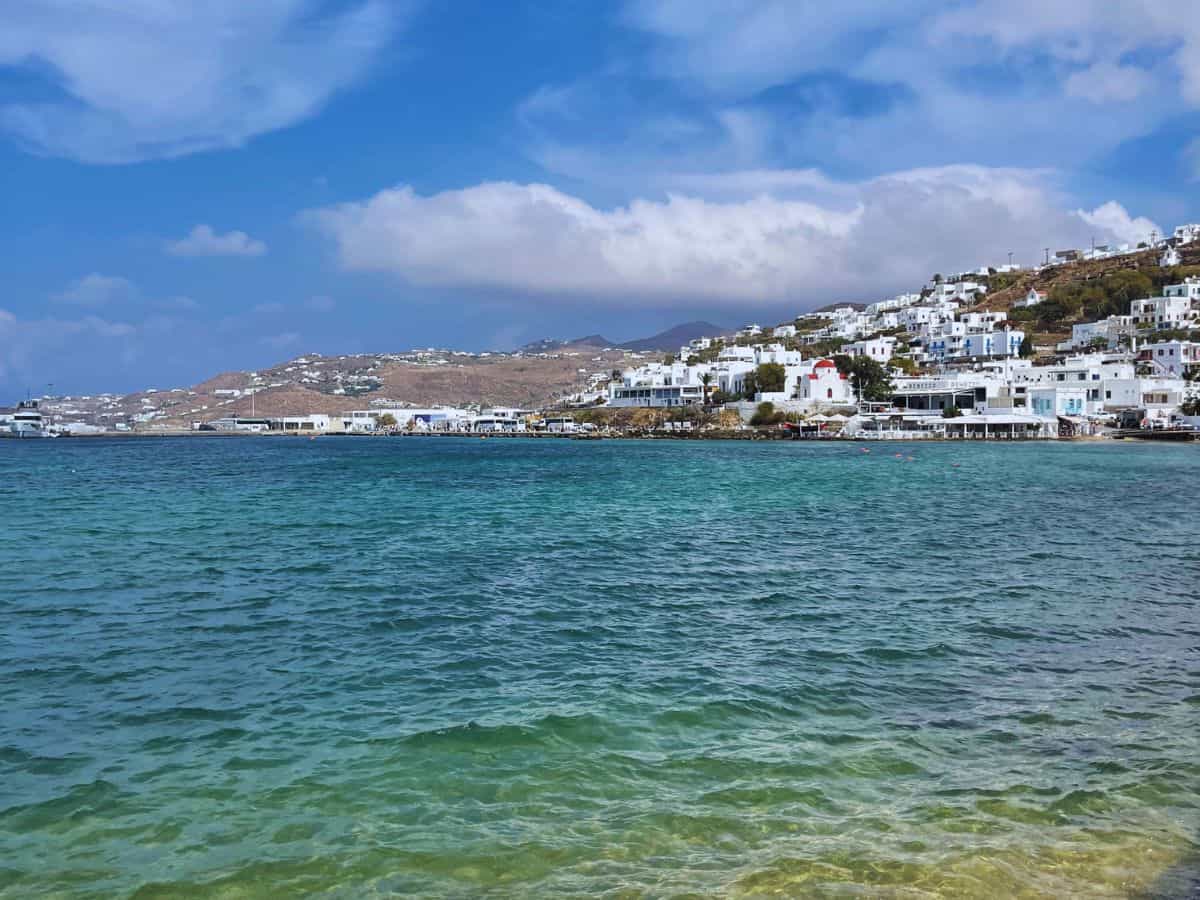 The tranquil azure waters of the Aegean Sea caress the white-washed buildings of Mykonos, with the island's hilly terrain rising in the background under a soft blue sky, embodying the quintessential beauty of Greek island architecture.