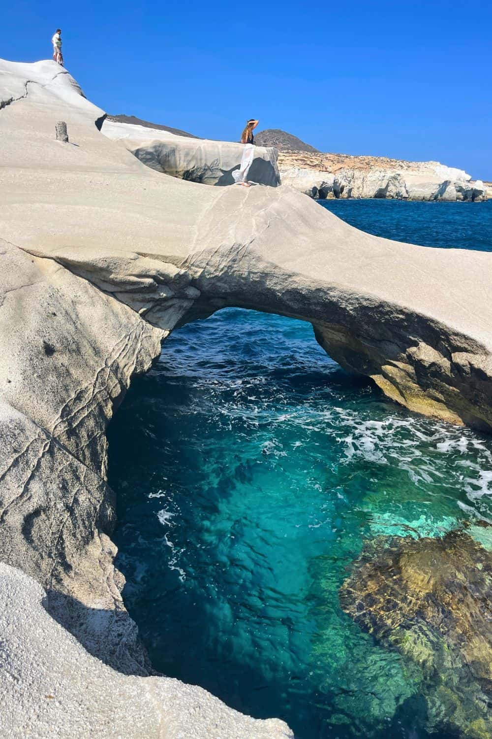 A striking photograph showcasing the geological beauty of Milos with its white rock formations creating a natural bridge over the clear turquoise waters. Two people are seen standing atop the formation, giving a sense of scale and adventure.