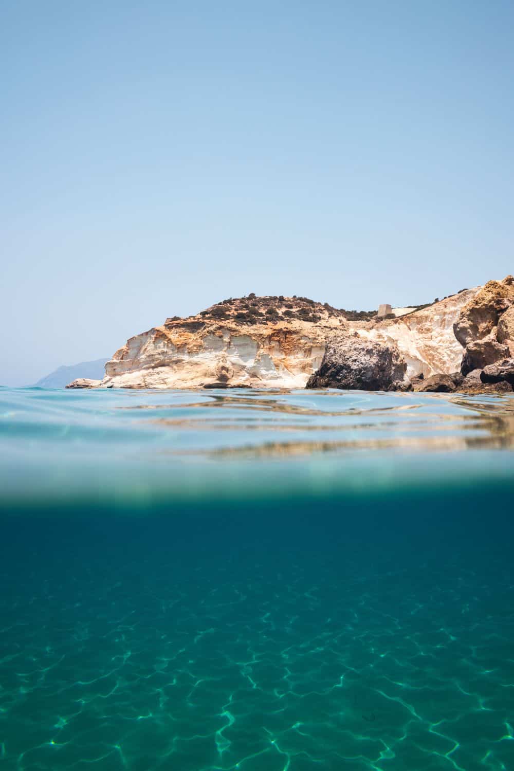 A reason Milos is worth visiting, the beaches. An underwater perspective half-submerged, showing the crystal-clear waters of Milos and the rocky landscape above, with a clear sky. This split view captures the dual beauty of Milos, both above and below the sea.