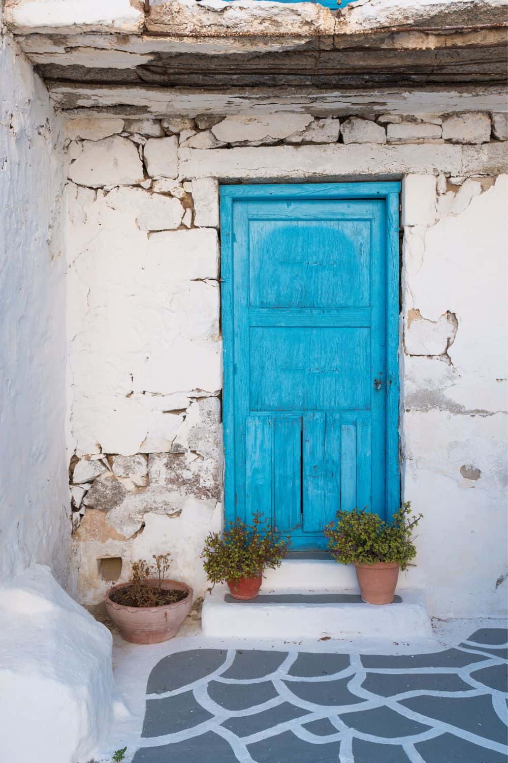 A textured image of a traditional Greek scene featuring a bright blue door set into a white-washed wall with peeling plaster. Potted plants sit on either side of the door, and the ground in front features a classic Greek design in grey and white. This image captures the charming architectural details found in Milos.