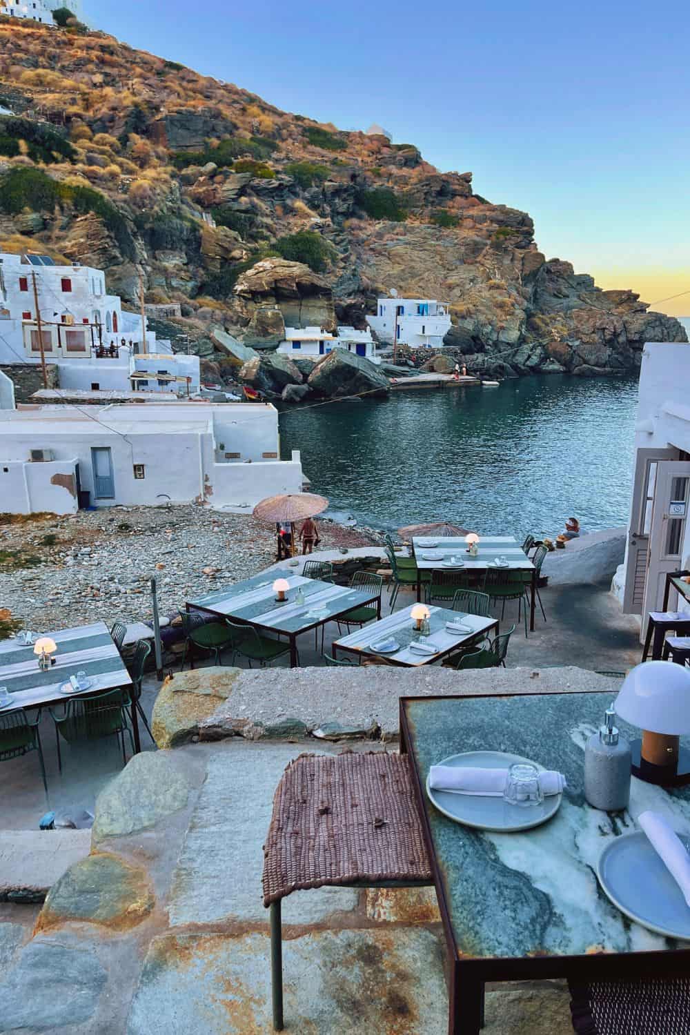 A picturesque outdoor dining setting in Sifnos overlooking a tranquil cove with azure waters. The tables are set with simple elegance against the backdrop of white-washed buildings and rugged cliffs, offering an immersive dining experience that prompts one to consider the charm of the island.
