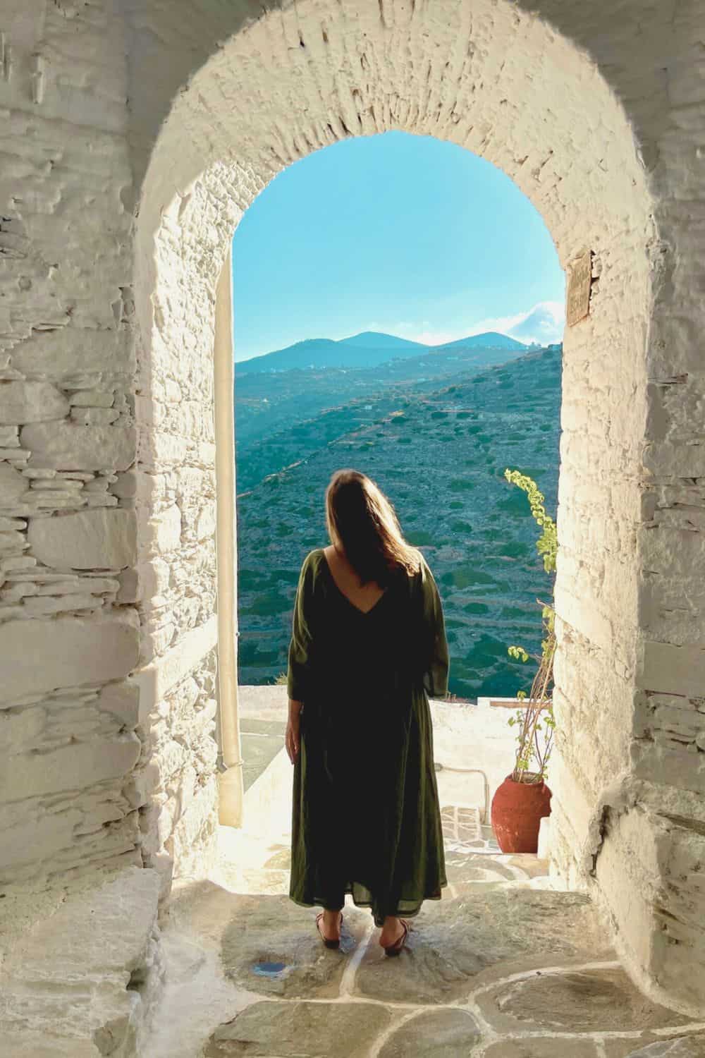 A woman stands in the shadow of an arched stone doorway, gazing out at the expansive landscape of Sifnos. The horizon is dotted with gentle hills under a clear blue sky, and the moment captures a sense of peaceful contemplation.