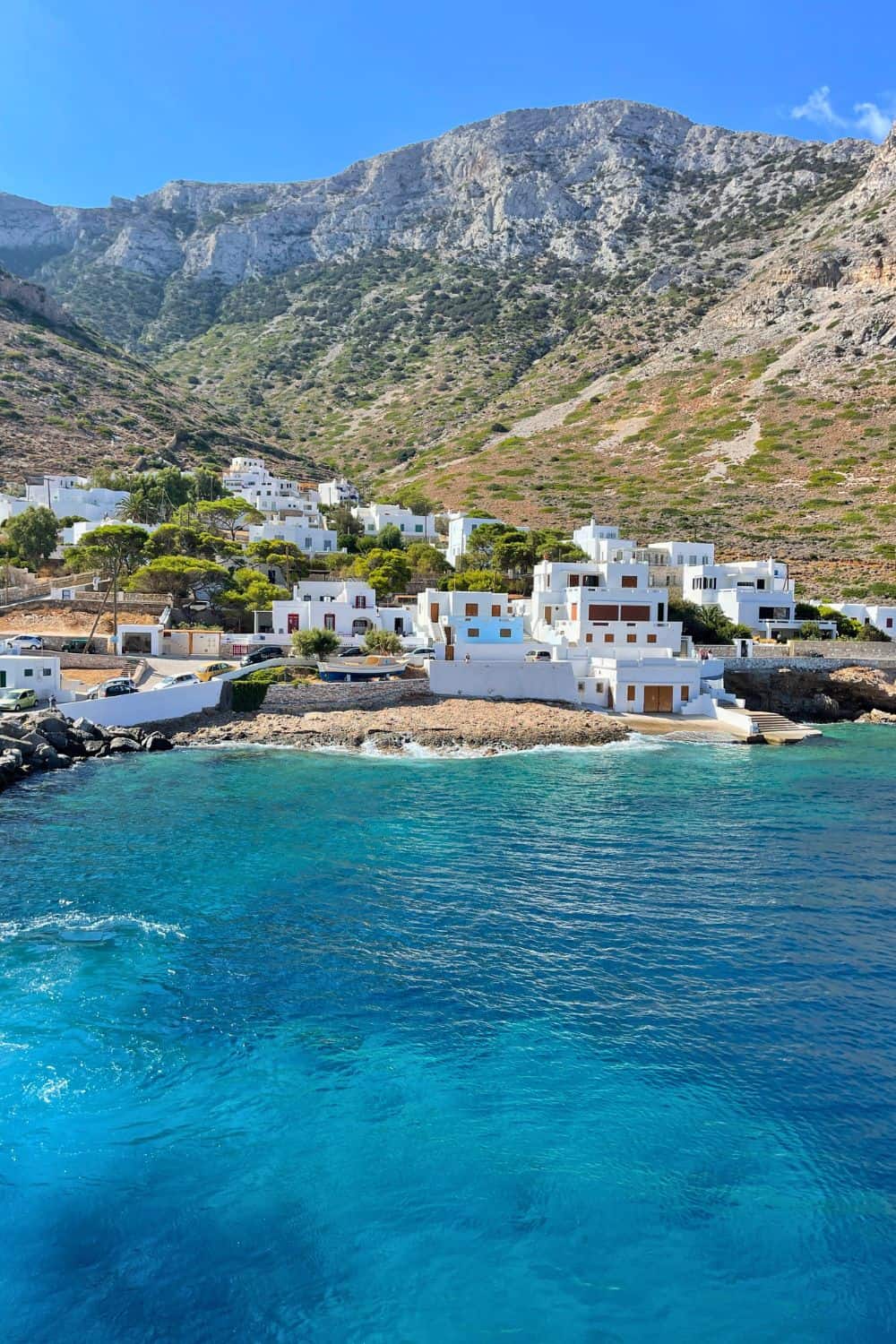 A tranquil coastal village in Sifnos nestled at the base of rugged hills, with whitewashed buildings reflecting the bright sunlight. The azure waters of the Aegean Sea gently kiss the rocky shoreline, inviting visitors to ponder the allure of the island.